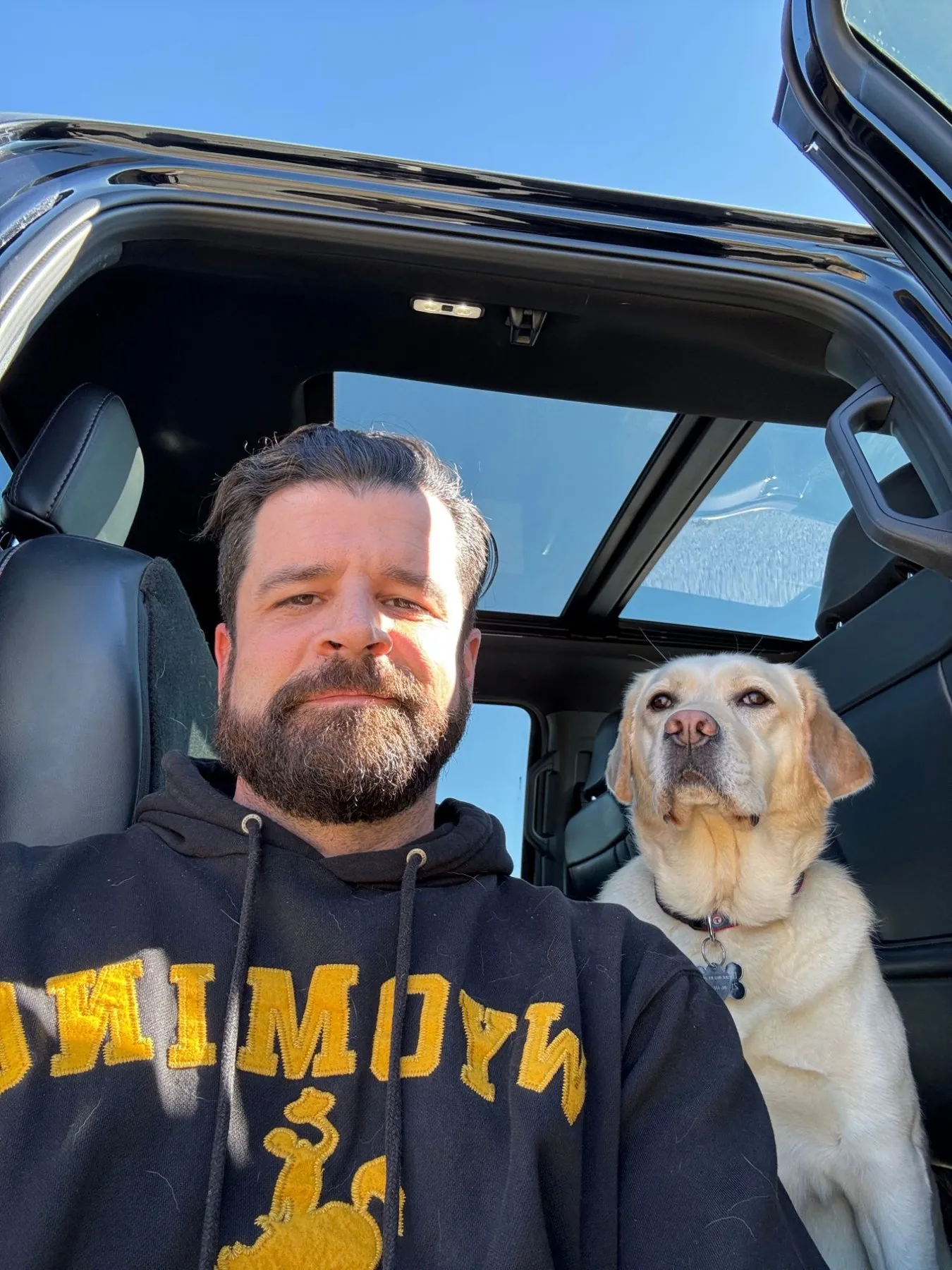 Drew, the host, smiling with a beard and a black hoodie, beside Ranger, a yellow Labrador, in a truck on a sunny day