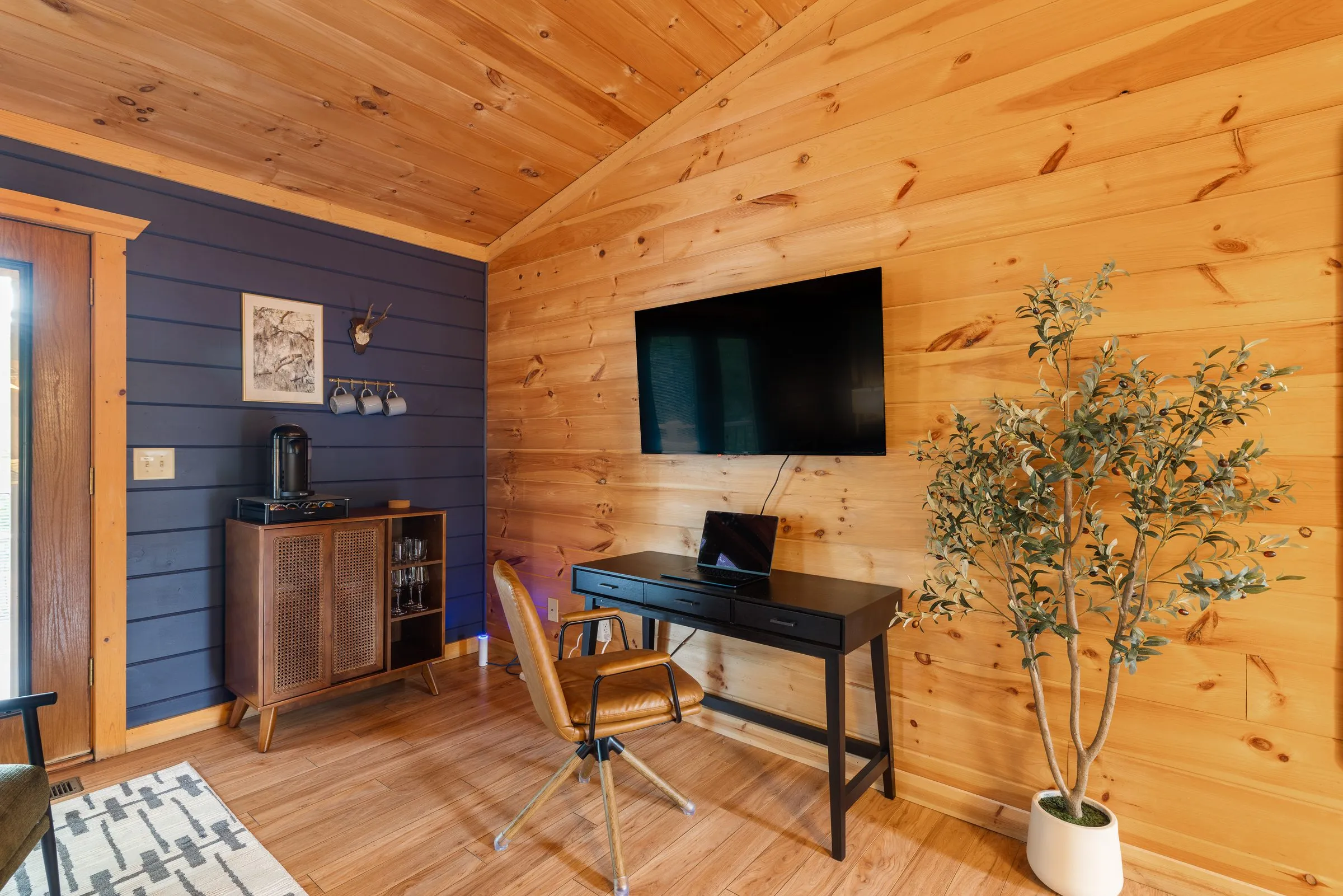 Primary suite sitting area with a desk, coffee bar cabinet, television and an olive tree under a vaulted wood ceiling