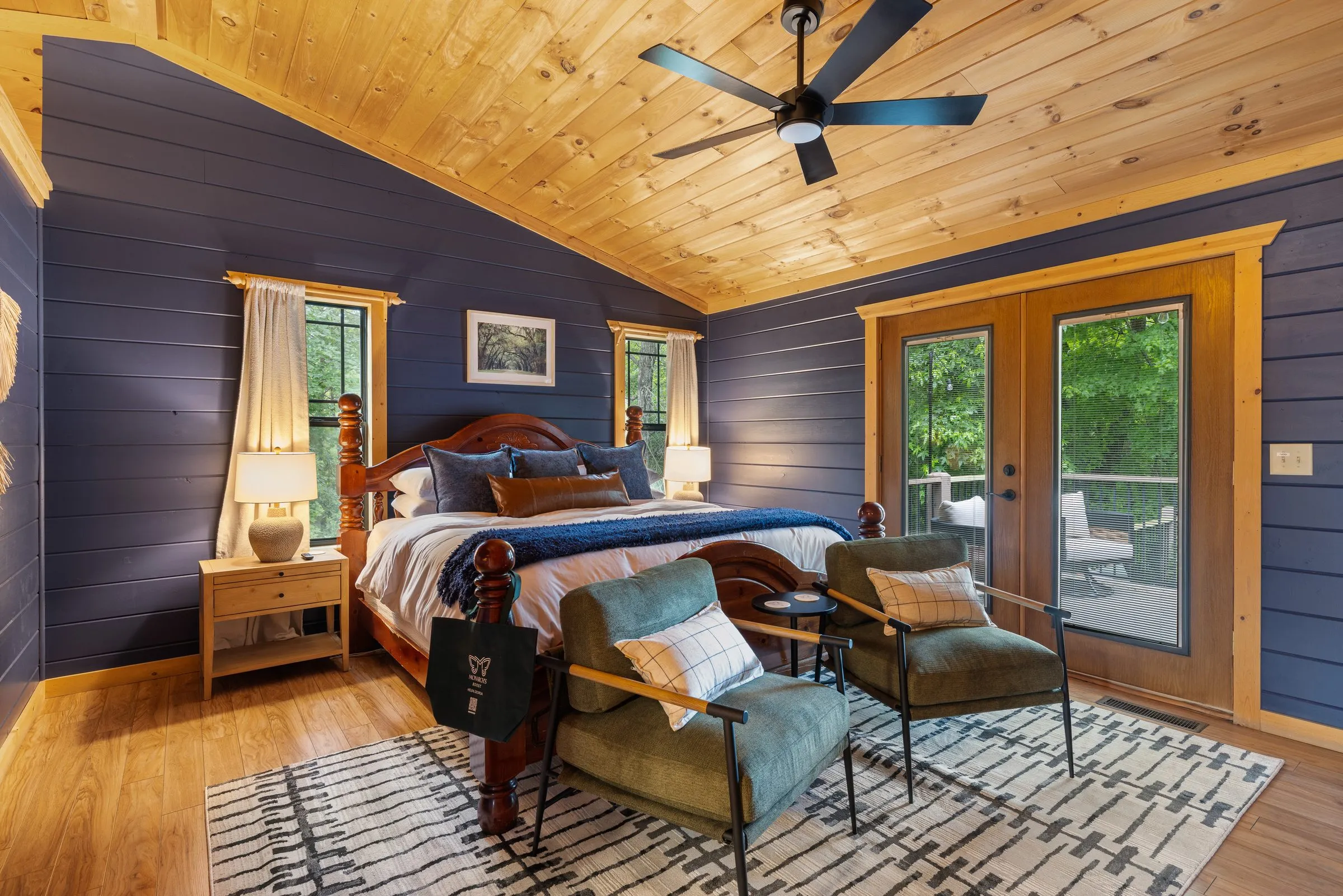 Primary suite with a carved four-poster king bed, navy walls, vaulted wood ceiling, green lounge chairs and French doors to the deck