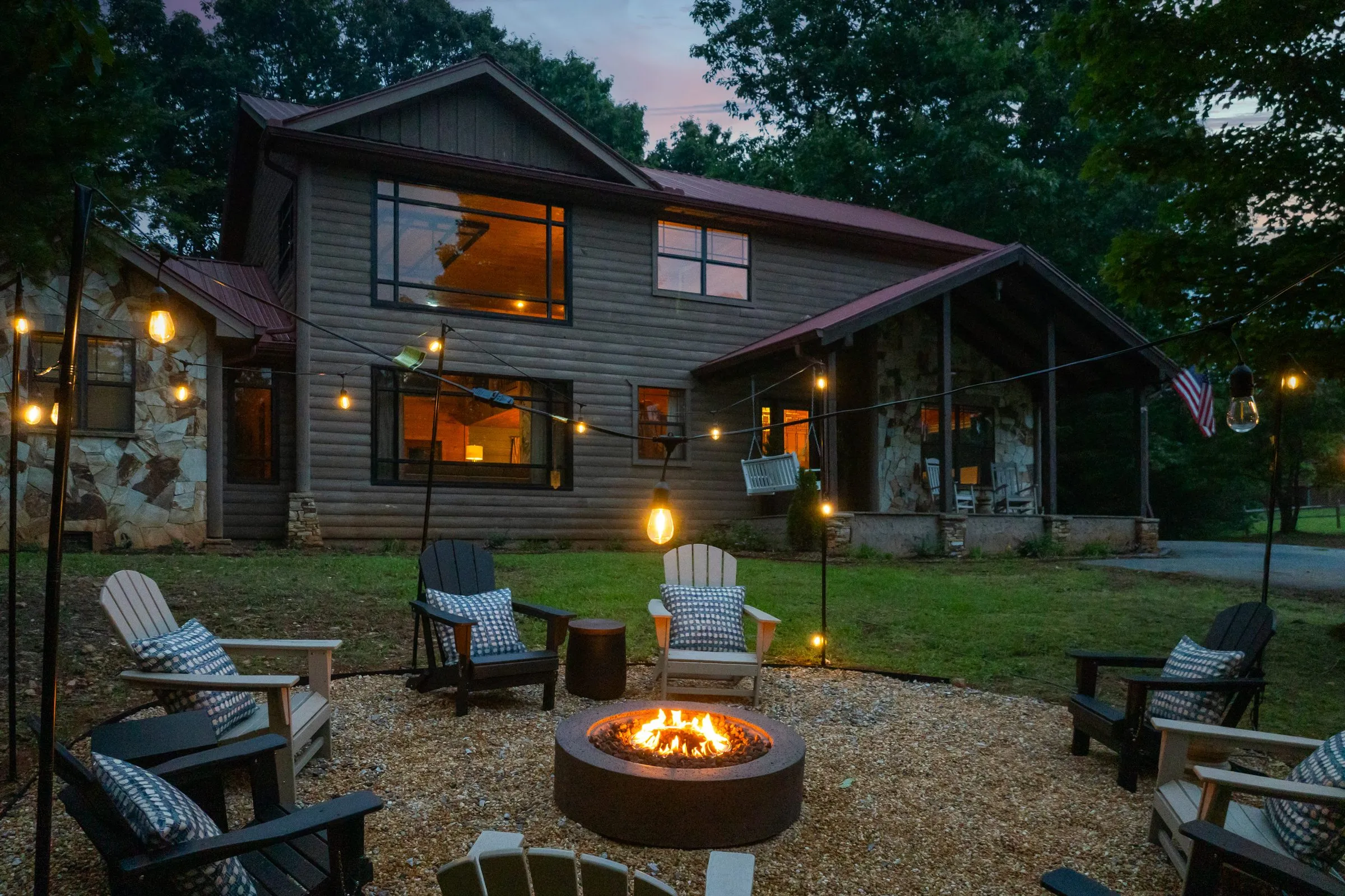 Lit fire pit ringed by Adirondack chairs under string lights at dusk, cabin behind