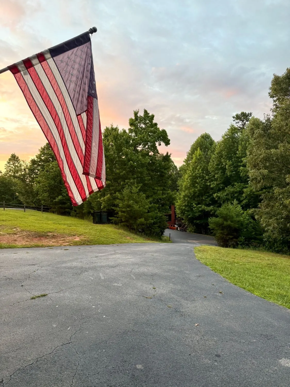 The American flag at the cabin's driveway against a pink and gold sunset sky, with tall trees beyond