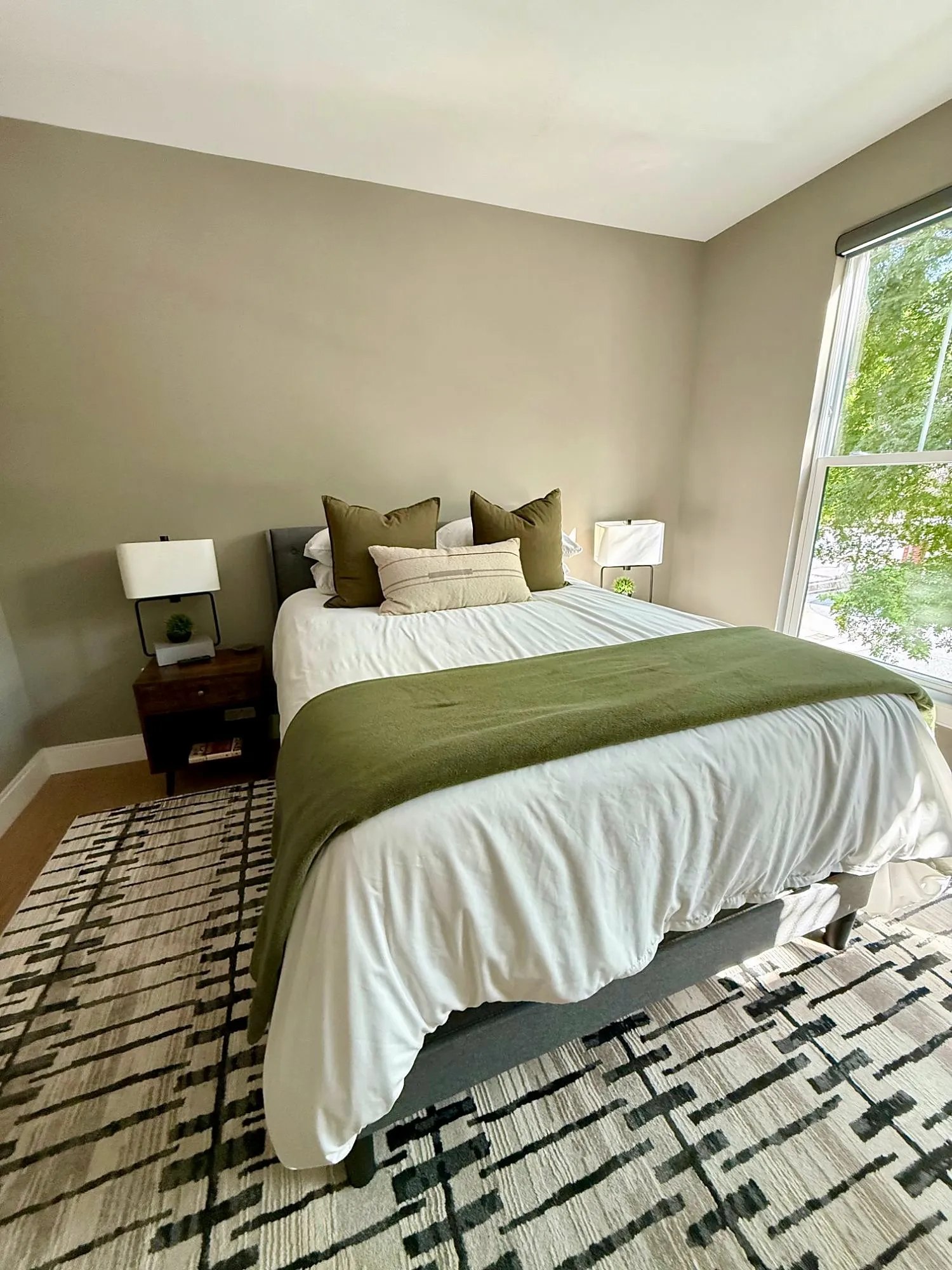 Bedroom with a queen bed, olive throw, two lamps and a graphic black and cream rug beside a tall window
