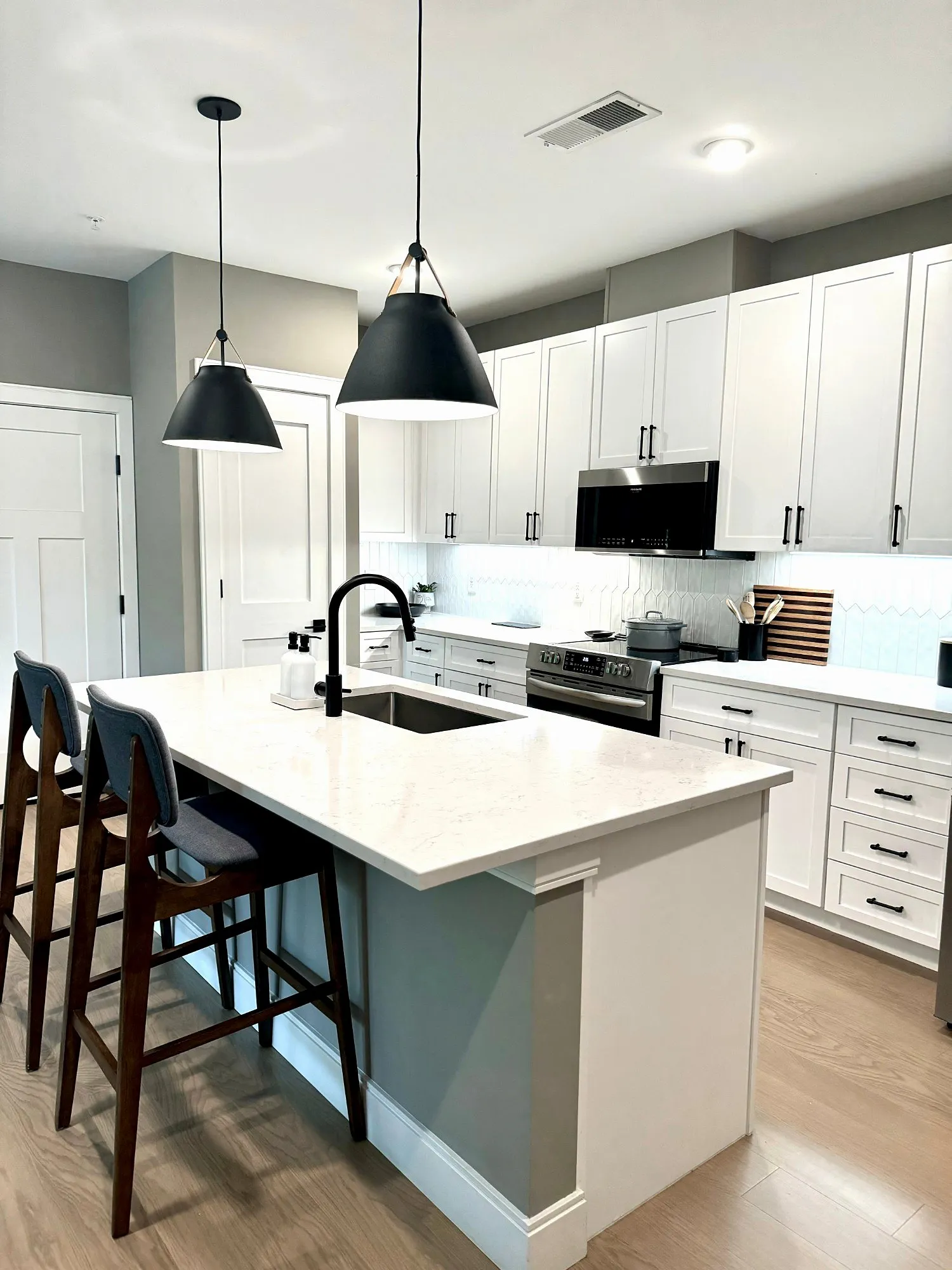 Kitchen with a white quartz island, black pendant lights, two bar stools, white cabinets and stainless appliances