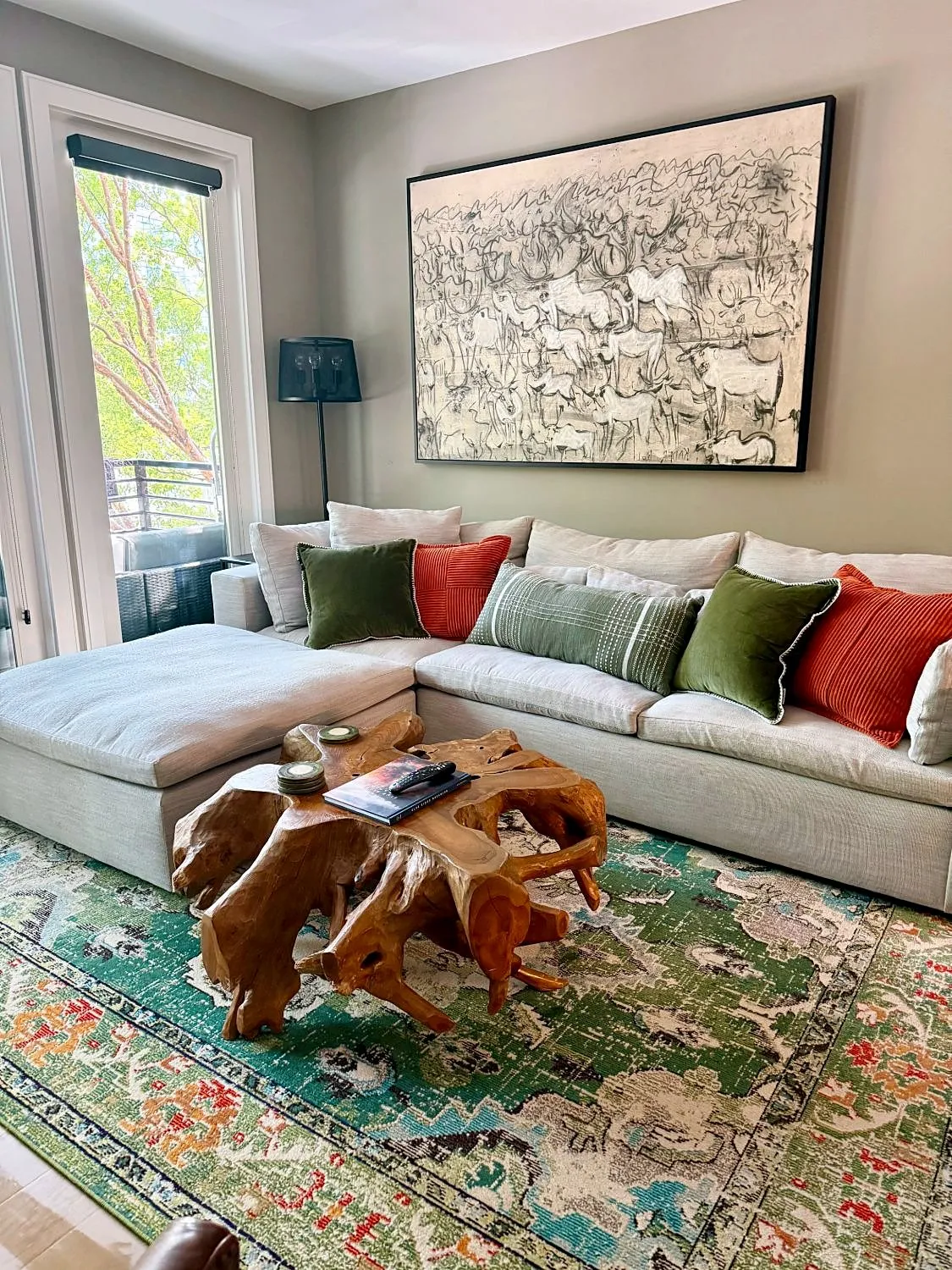 Sofa with green and rust pillows beneath a large charcoal drawing, with a teak root coffee table on a green rug