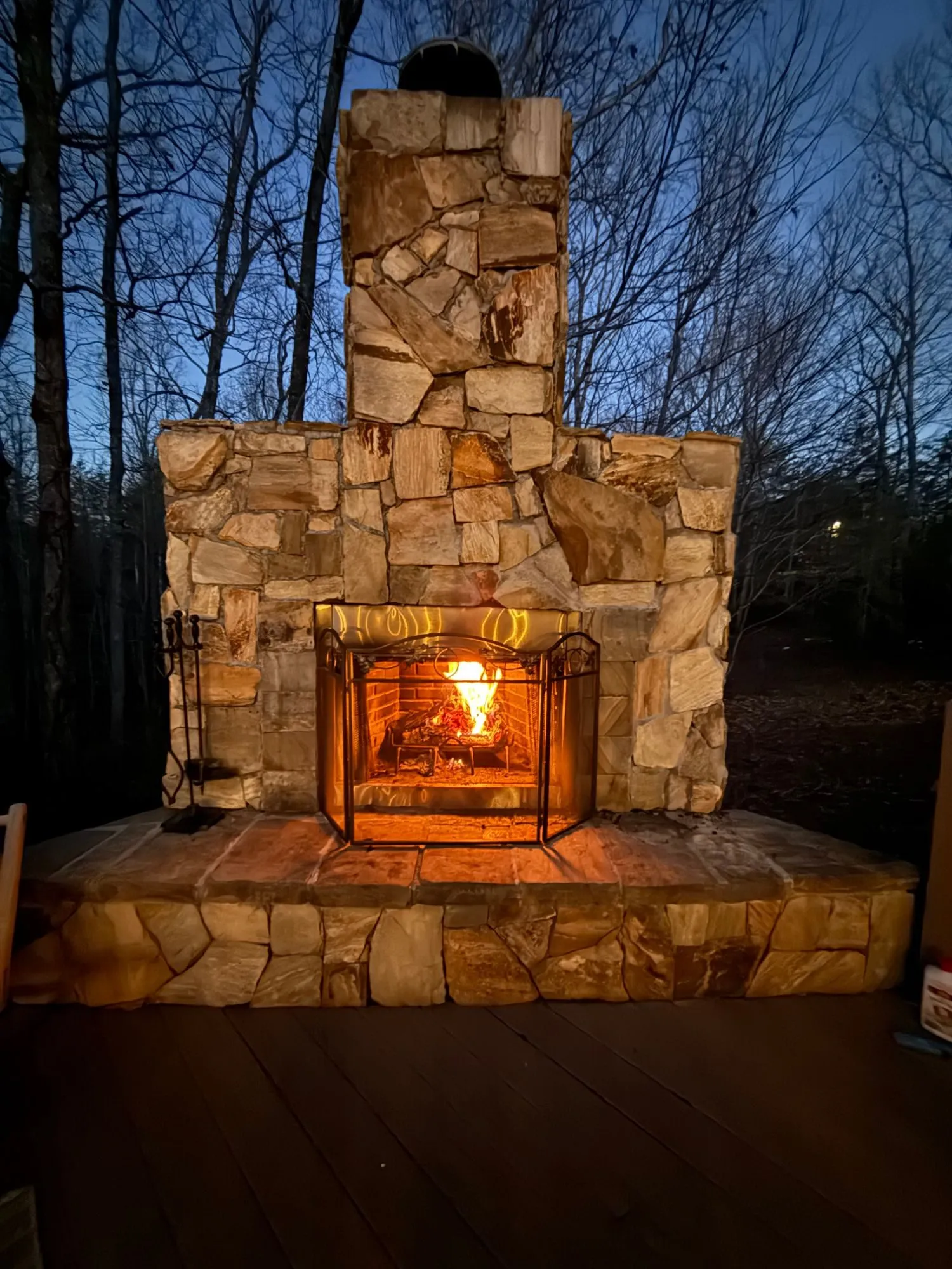Outdoor stone fireplace with a wood fire burning at dusk on the back deck, with bare trees and a blue evening sky behind