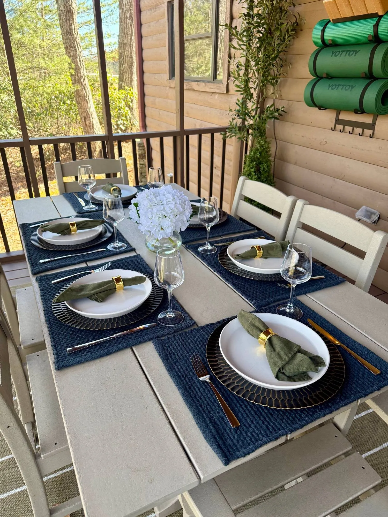 Screened porch dining table set for six with navy placemats, olive napkins with gold rings, wine glasses and white hydrangeas